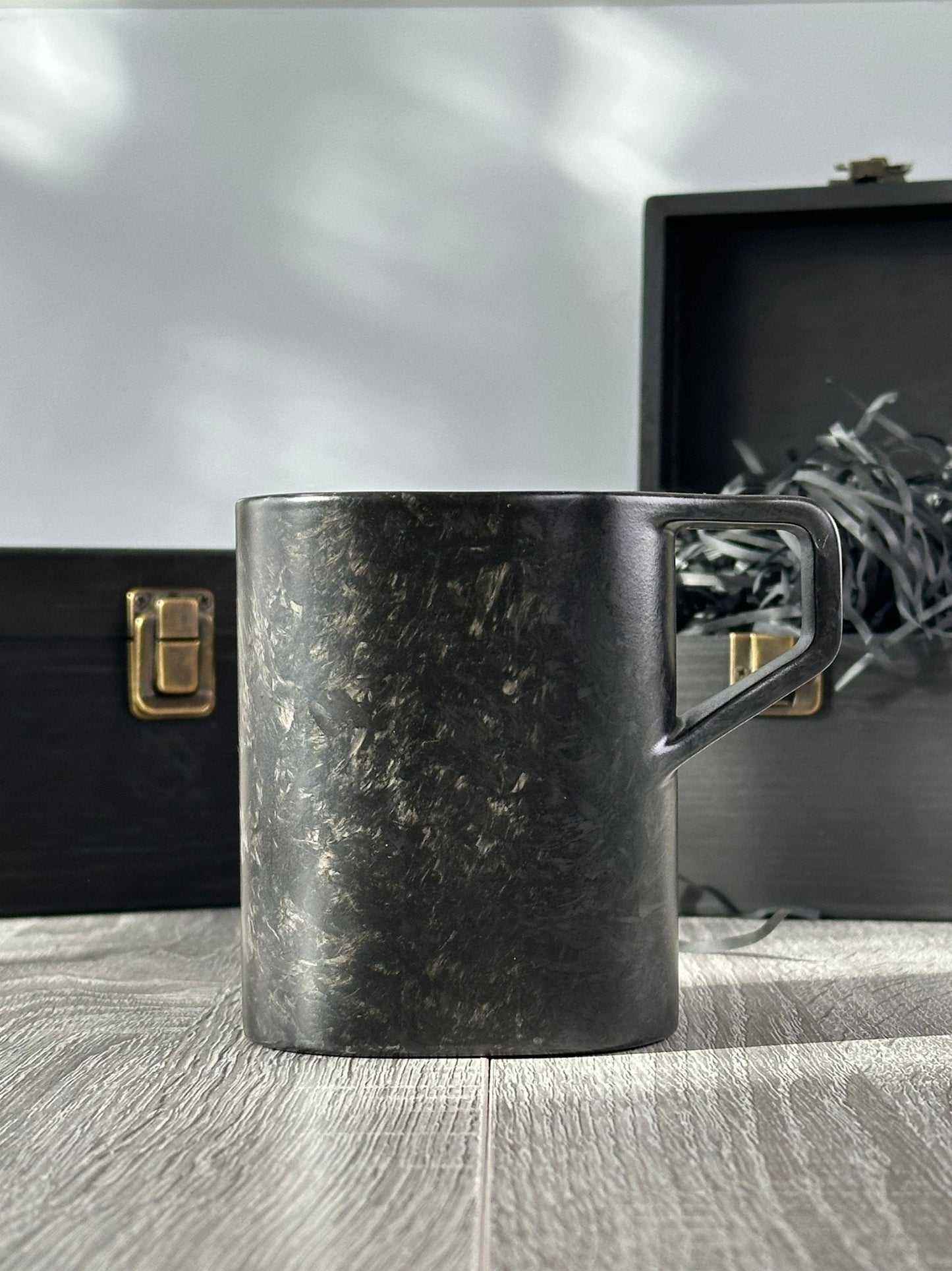 Side profile of virgin carbon fiber mug with open and closed wooden boxes behind